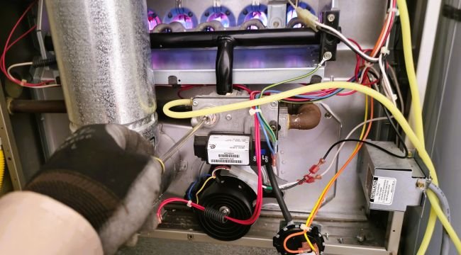 Technician repairing a gas furnace burner assembly with active blue flames and exposed wiring.